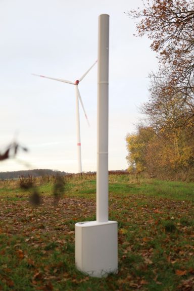 Weißer Mast im Vordergrund mit Windrad im Hintergrund, umgeben von Bäumen.